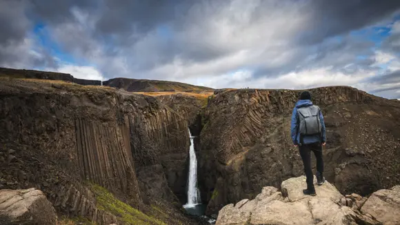 A hiker standing at the summit of a rocky mountain in Iceland admiring the view featuring a waterfall