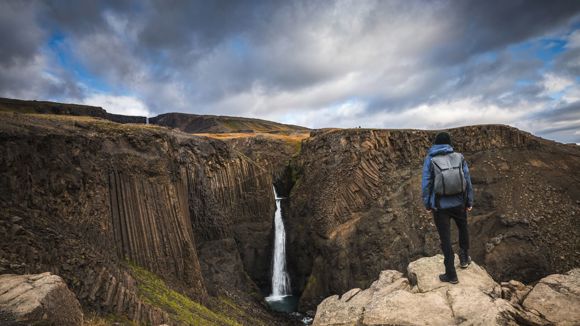 A hiker standing at the summit of a rocky mountain in Iceland admiring the view featuring a waterfall