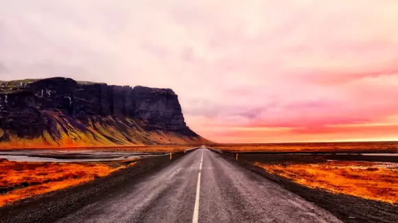 A highway in Iceland with rocky mountains and pink skies in the background.