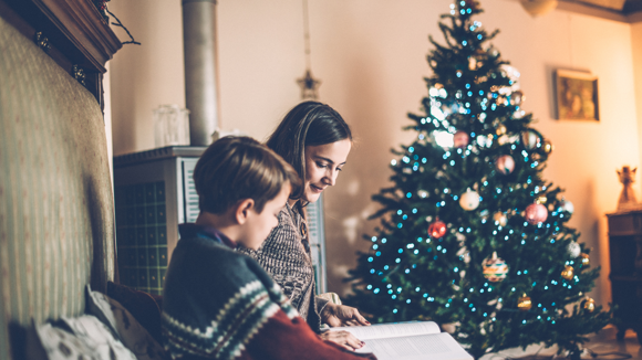 A woman reading a book to a boy with a Christmas tree in the background.