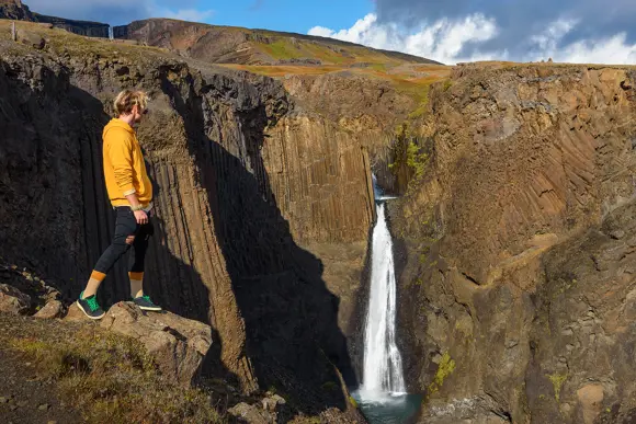 Hiker wearing yellow looking back at Litlanesfoss Waterfall.